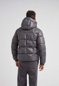 Grå pufferjakke med hætte, med et quiltet design, elastiske manchetter og et blåt mærke på ryggen. Glat, blank overflade.