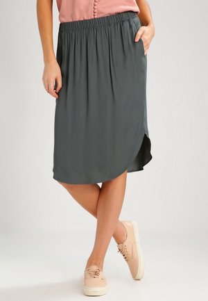 Donna che indossa una gonna midi grigio scuro con cintura elastica e orlo arrotondato, una maglietta rosa pallido e sneakers rosa chiaro, in piedi con le gambe incrociate.
