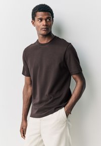 T-shirt marron à manches courtes en tissu doux avec un col rond, associé à un pantalon beige clair. Le modèle est posé devant un fond neutre.