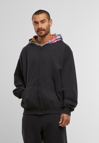 Mann mit kurzen Haaren und Bart, der einen schwarzen Reißverschluss-Hoodie mit buntem gemustertem Kapuzenfutter und passenden schwarzen Hosen trägt, steht vor einem grauen Hintergrund.