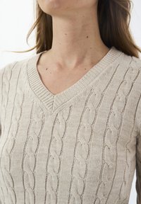 Vrouw in een beige kabelgebreide V-hals trui, met zichtbare nek en bovenborst, en lange bruin haar over haar schouders.
