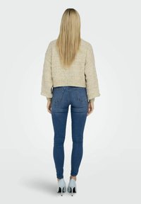 Pull en maille beige coupé, avec des manches larges, associé à un jean bleu ajusté et des chaussures à talons hauts bleu clair. Vue de dos.