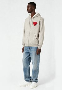 Sweat à capuche zippé gris avec une doublure en polaire blanche, orné d'un graphique de cœur rouge avec un design de main. Associé à un jean en denim bleu clair et des baskets blanches.