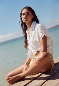 Hvid linnedskjorte bundet i taljen, kombineret med et gult og hvidt ternet skørt. Sidder på en trækaj ved vandet med en klar blå himmel.