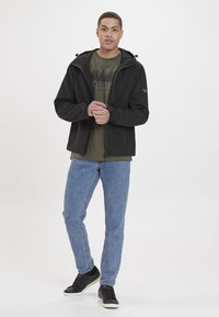 Giacca softshell nera con zip frontale, cappuccio regolabile e tasche doppie, indossata sopra una t-shirt grafica verde e jeans blu.