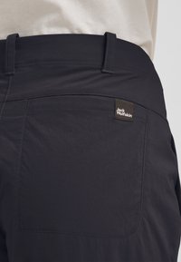 Dunkelgraue Hose mit einer Gesäßtasche, die mit einem kleinen braunen Markenetikett versehen ist. Das Material hat eine glatte Textur und einen dezenten Glanz.
