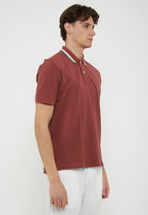 Giovane uomo in piedi di profilo che indossa una polo color ruggine con colletto a righe e pantaloni bianchi su sfondo neutro.