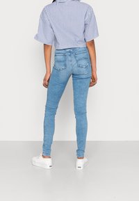 Mujer que lleva jeans ajustados de color azul claro, zapatillas de plataforma blancas y una camisa de rayas azules y blancas de manga corta, de pie con la espalda hacia la cámara.
