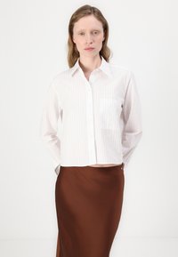 Hvit og lys rosa stripete skjorte med knapp, med en brystlomme, kombinert med et brunt satengskjørt. Stoffet ser jevnt og litt teksturert ut.