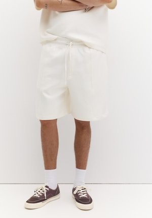 Person trägt weiße Shorts mit Kordelzug, weiße Socken und dunkelbraune Flach-Turnschuhe und steht vor einem schlichten weißen Hintergrund.