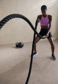 Cordes de combat noires, une silhouette athlétique portant un haut sans manches violet et un short noir, les utilisant sur un fond de teinte neutre.
