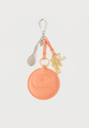 Llavero redondo de cuero color melocotón con el logo de Lacoste en relieve, unido a un charm dorado de cocodrilo, un charm con la letra "L" y un charm plateado de raqueta de tenis.