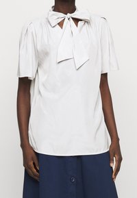 Světlá šedá blůza s krátkými nabíranými rukávy a mašlí na výstřihu; hladká textura a volný střih.