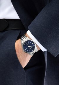 Montre en acier inoxydable argenté avec un cadran bleu marine, des index argentés et une fonction date, portée sur un poignet sous une veste de costume sombre.