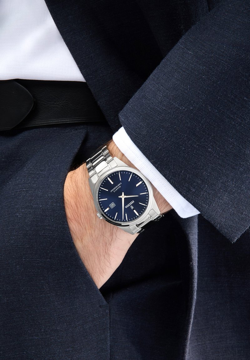 Montre en acier inoxydable argenté avec un cadran bleu marine, des index argentés et une fonction date, portée sur un poignet sous une veste de costume sombre.