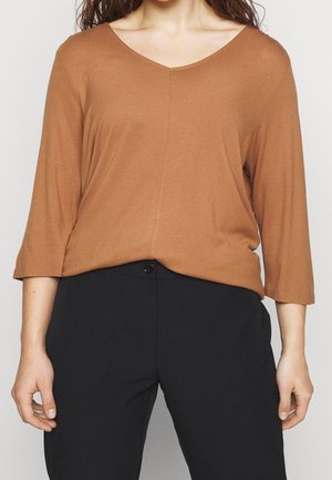 Femme portant un haut marron à encolure en V, manches trois-quarts et un pantalon noir taille haute avec fermeture par bouton.
