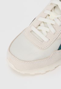 Sneaker beige e bianca con lacci bianchi, realizzata in materiale scamosciato e mesh, e una porzione di una striscia verde scuro sul lato.