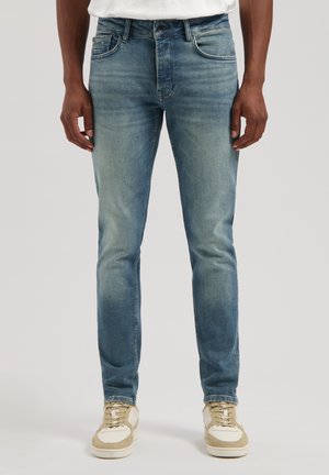Light blue denim jeans with a slim fit, subtle whiskering, and five-pocket design. Paired with beige and white low-top sneakers.