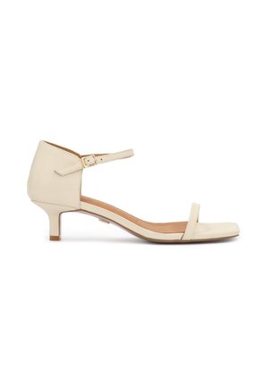 Beżowy sandał na niskim obcasie z paskiem wokół kostki, kwadratowym noskiem i pojedynczym paskiem na palce, pokazany z boku na białym tle.