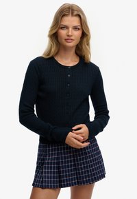 Mørkeblå strikket cardigan med tekstureret kabelmønster, rund halsudskæring og knaplukning foran. Kombineret med et plisseret ternet nederdel.