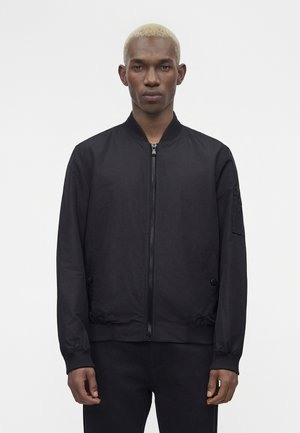 Jeune homme aux cheveux blonds courts, portant une veste bomber noire zippée et un pantalon noir, debout devant un fond blanc uni.