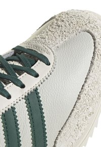 Sneaker avec un dessus en cuir blanc lisse orné d'accents rayés en zigzag verts, complété par un panneau en daim gris texturé. Lacets verts.