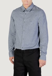 Camicia a maniche lunghe con bottoni in un tessuto blu-grigio testurizzato. Presenta un colletto classico, bottoni frontali e un logo discreto sul petto. Pantaloni neri.