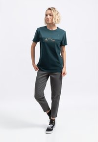 Teal katoenen t-shirt met een geprinte ontwerp op de borst. Grijze broek met een gladde textuur. Zwarte sneakers met witte veters.