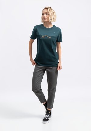 T-shirt en coton teal avec un design imprimé sur la poitrine. Pantalons gris avec une texture lisse. Baskets noires avec des lacets blancs.