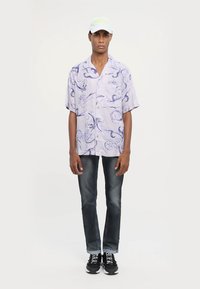 Joven vestido con una gorra blanca, una camisa de manga corta de botón en un tono púrpura claro con patrones en espiral, jeans negros desgastados y zapatillas negras.
