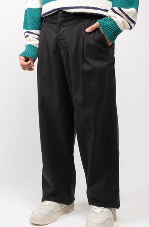 Schwarze Baumwollhose mit lockerer Passform, ausgestattet mit Falten und seitlichen Taschen. Kombiniert mit weißen Sportschuhen und einem mehrfarbigen Pullover.