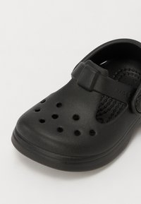 Zueco de espuma negra con puntera cerrada, múltiples agujeros de ventilación y una correa ajustable para un ajuste seguro. Textura suave y diseño ligero.