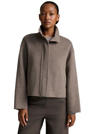 Donna in piedi che indossa un cappotto corto in lana color taupe con collo alto e maniche lunghe, abbinato a pantaloni marrone scuro.