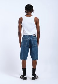 Shorts de mezclilla en azul medio con un ajuste relajado, bajos deshilachados y bolsillos traseros. Combinados con una camiseta sin mangas blanca y calcetines negros.