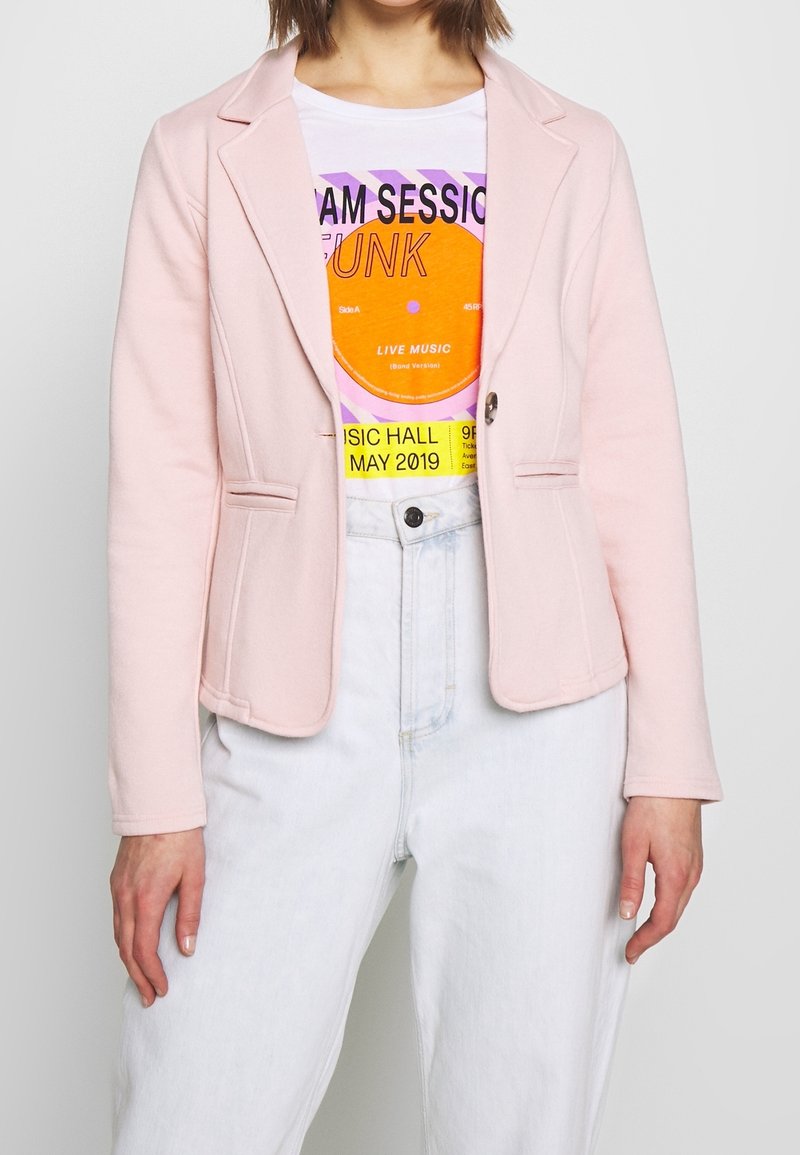 Ljusrosa kavaj med skräddarsydd design, med två framfickor, ensamt knäppning och mjukt texturerat tyg, buren över en grafisk T-shirt.