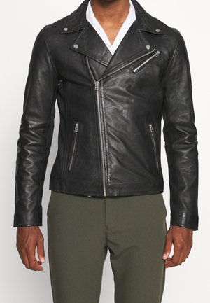 Homme portant une veste de motard en cuir noir avec des fermetures éclair argentées et un pantalon vert olive sur un fond uni.