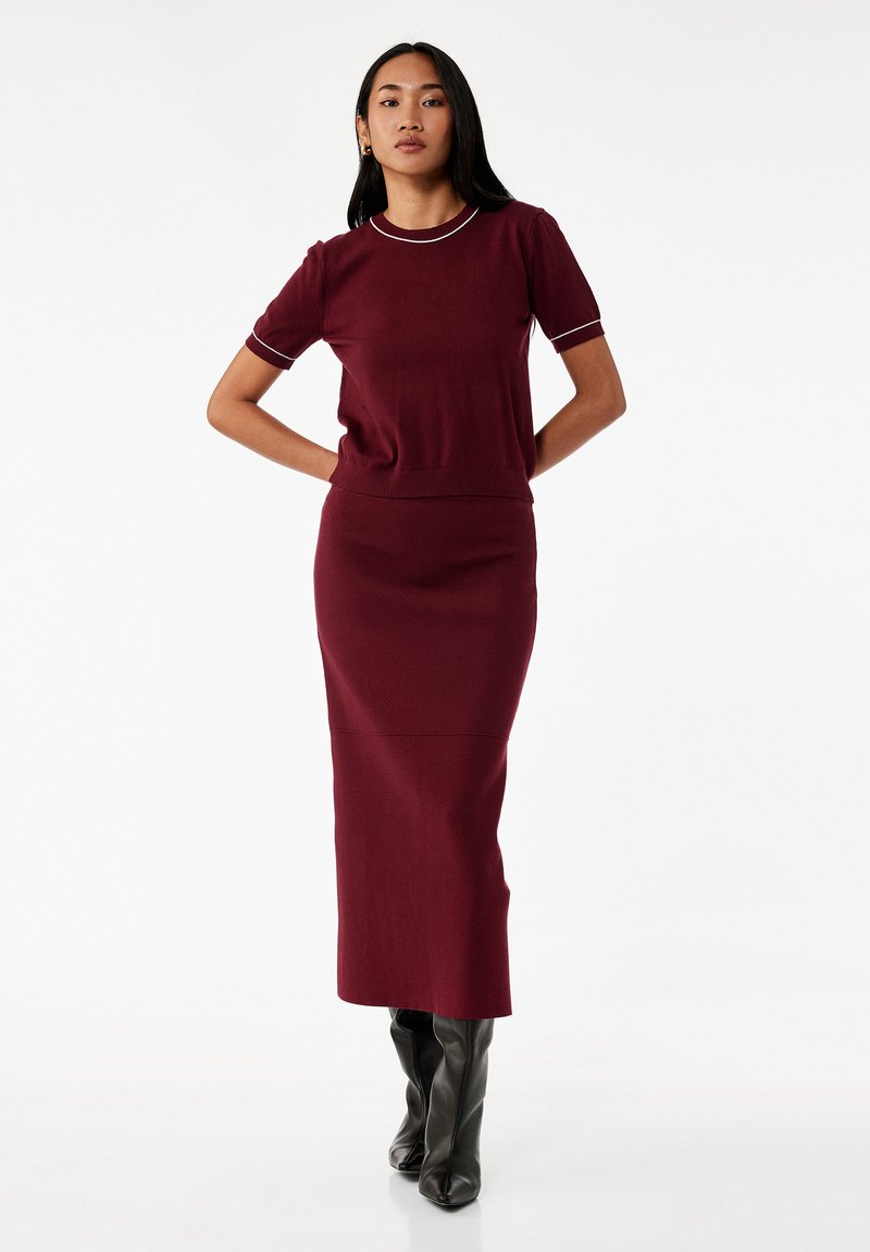 Kobieta stoi z rękami za plecami, ubrana w bordowy, krótki top i dopasowaną, długą spódnicę oraz czarne botki do kolan.