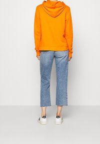 Felpa arancione con cappuccio e tasca a marsupio, abbinata a jeans a gamba larga blu chiaro con orli sfilacciati. Indossata con sneaker bianche.
