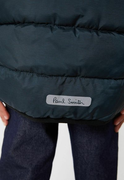 Pildīta tumši zaļa veste ar horizontālu šuvju rakstu un pelēku etiķeti, uz kuras redzams "Paul Smith", apakšmalā. Valkāta virs tumšajām džinsu biksēm.