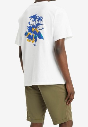 Mężczyzna w białej koszulce z niebiesko-żółtym motywem kwiatów i palm, ubrany w oliwkowozielone szorty, widziany od tyłu.