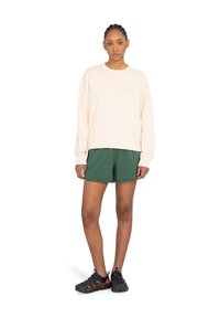 Cremefarbener Pullover mit gerippten Bündchen, kombiniert mit grünen Shorts und schwarzen Sportenschuhen. Der Pullover hat ein kleines Markenlogo auf der Brust.