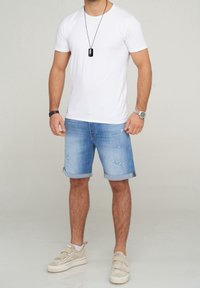 Camiseta blanca, pantalones cortos de mezclilla azul con dobladillos enrollados y detalles desgastados, zapatillas beige con correas, collar de etiqueta negra.