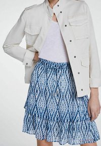 Veste en denim blanc avec deux poches poitrine, associée à une jupe à niveaux bleu et blanc à motifs, avec une taille à cordon et des accents en argent.