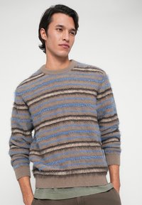 Young man with short dark hair wearing a fuzzy striped sweater in beige, blue, and brown, standing with hands in pockets against white background.