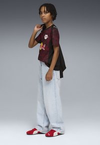 Czerwona i czarna koszulka z logo i geometrycznym wzorem, zestawiona z jasnoniebieskimi szerokimi dżinsami i czerwono-białymi trampkami. Minimalistyczny design.