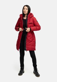 Red, insulated parka with a hood and adjustable belt, featuring large front pockets and a zipper closure. Worn over black attire and boots.