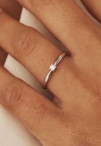 Silberring mit einem gebogenen Band, das einen einzelnen runden Diamanten in zarten Krappen fasst. Sanfte Textur, minimalistisches Design.