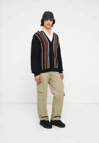 Sort cardigan med lodrette striber i rød, hvid og tan; beige cargo-bukser; sorte sneakers; og en sort kasket. Model står foran en hvid baggrund.