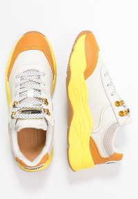 Valkoiset ja keltaiset urheilukengät oransseilla yksityiskohdilla, verkkomateriaaleilla ja metallisilla nauhoilla. Kenkien paksu kumipohja ja teksturoitu materiaali tarjoavat mukavuutta ja tukea.