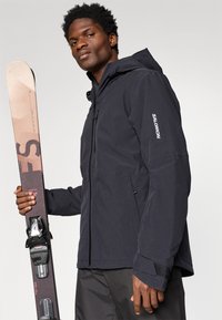 Homem com um casaco preto da Salomon segurando um único esqui de freestyle com fixações, de pé contra um fundo claro e liso.